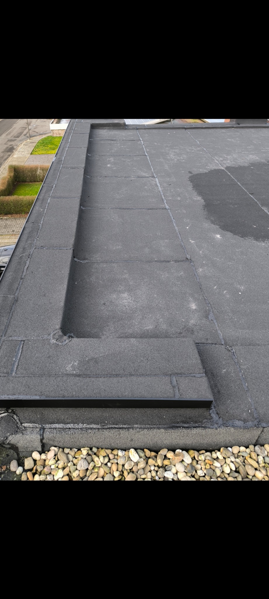 Płaski dach pokryty papą z widocznym obrzeżem z kamieni, zabezpieczający przed wodą. Fragment dachu z kałużą. Widok z góry na budynek.