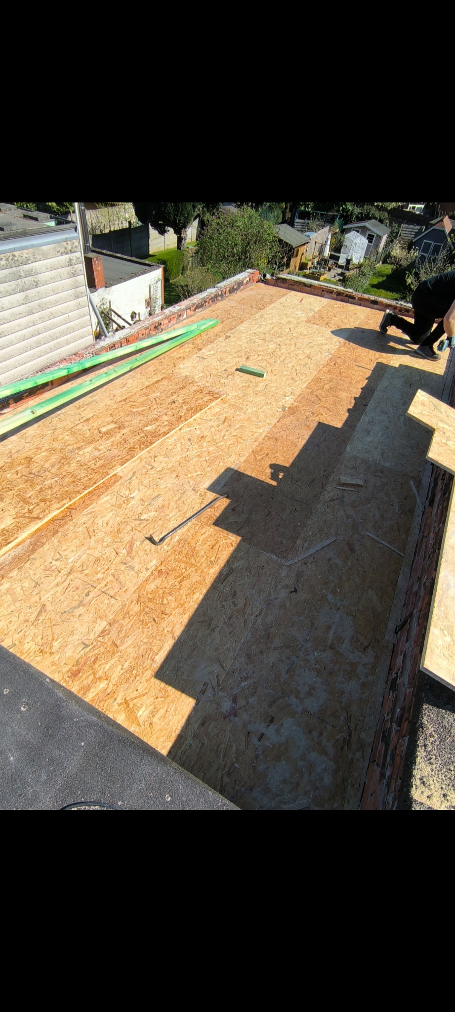 Montaż płyt OSB na płaskim dachu z ceglanym obrzeżem. Widoczne zielone listwy i częściowo ułożone płyty, tworzące wzór cienia. Prace dekarskie w toku.