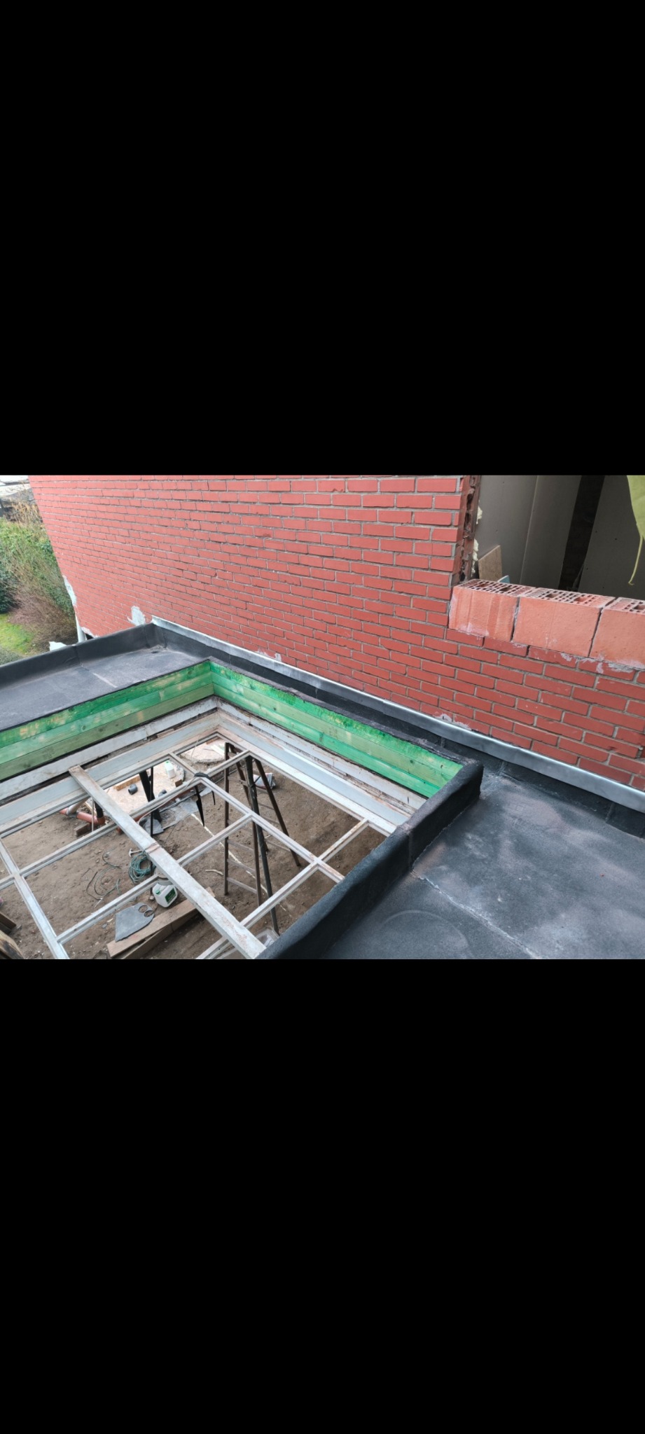 Dach płaski w trakcie naprawy. Widoczna konstrukcja nośna i fragment murowanej ściany z czerwonej cegły. Elementy zielonej impregnacji drewna.