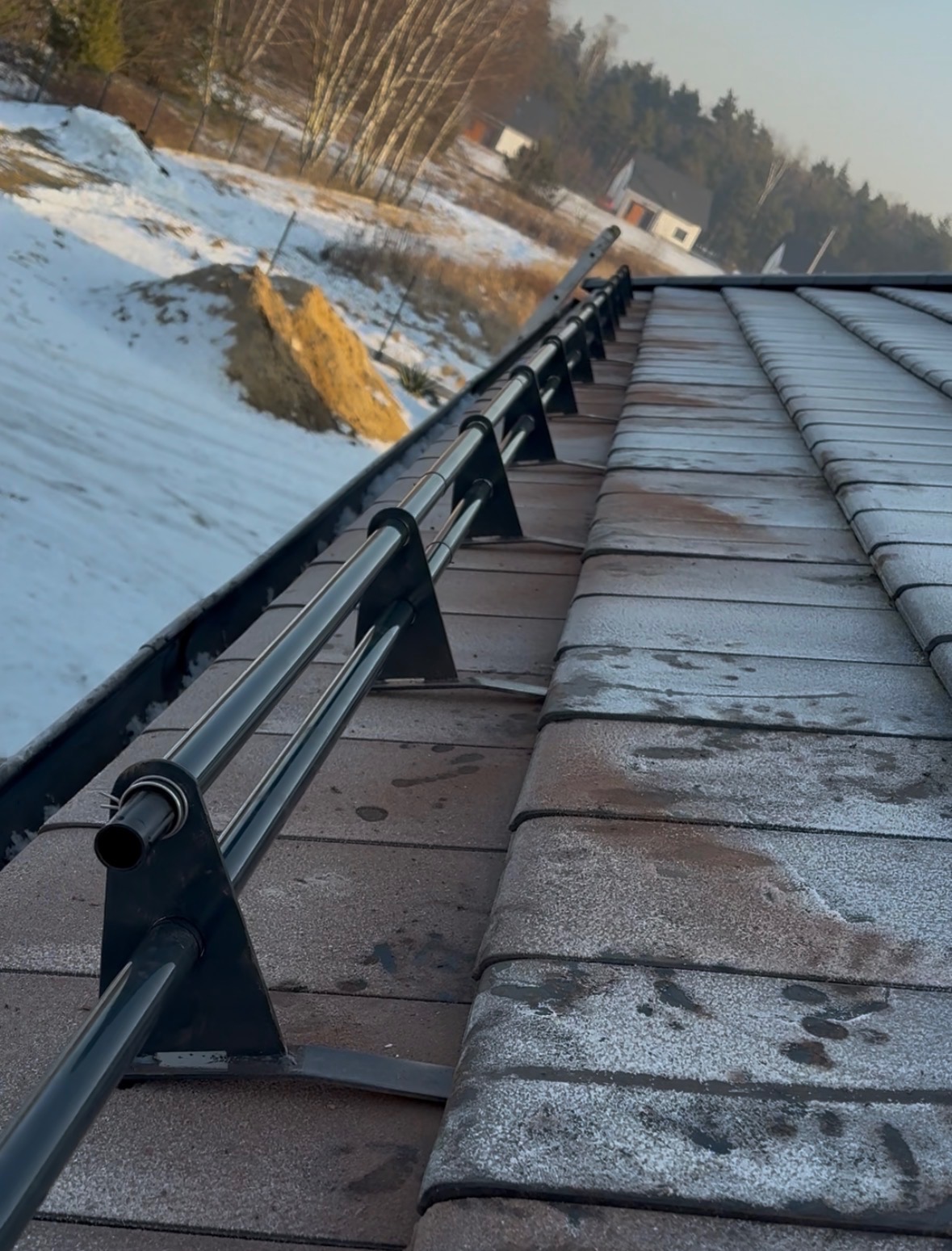 Zimowy krajobraz z widokiem na brązowy dach pokryty śniegiem i zamontowane czarne zabezpieczenia przeciwśniegowe. W tle drzewa i budynek.
