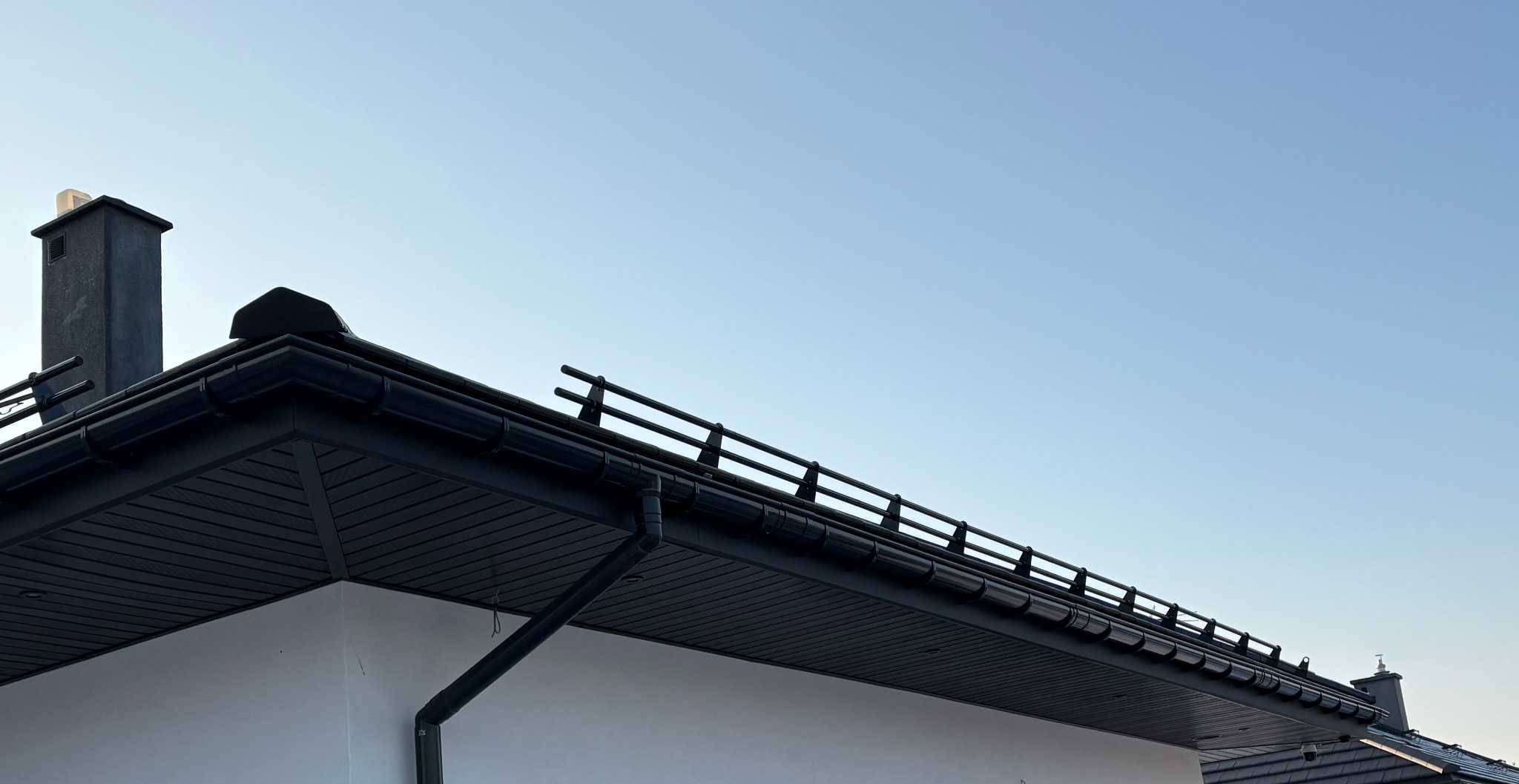 Fragment dachu z czarną blachodachówką, rynnami i kominem na tle błękitnego nieba. Widoczny fragment białej ściany budynku.