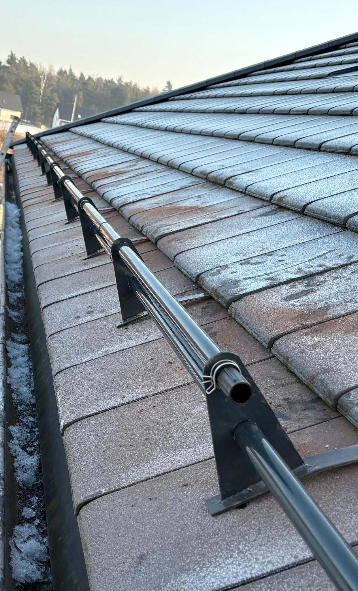 Szary dach pokryty dachówką ceramiczną z zamontowanym systemem odśnieżania w postaci metalowej rury, widoczny fragment rynny z resztkami śniegu.