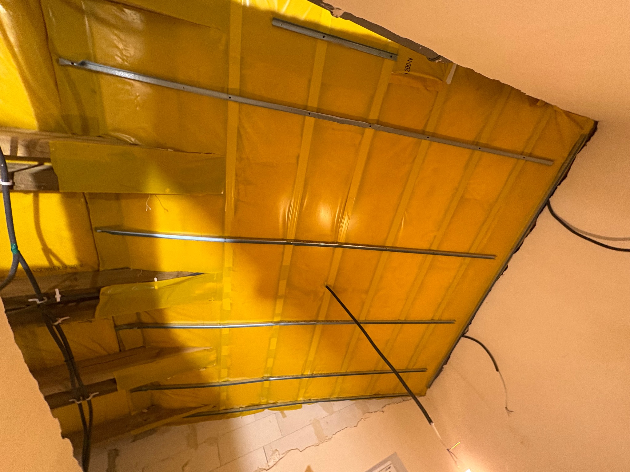 Żółta folia izolacyjna poddasza zamontowana na drewnianej konstrukcji z metalowymi profilami, widok z dołu, etap prac ociepleniowych.