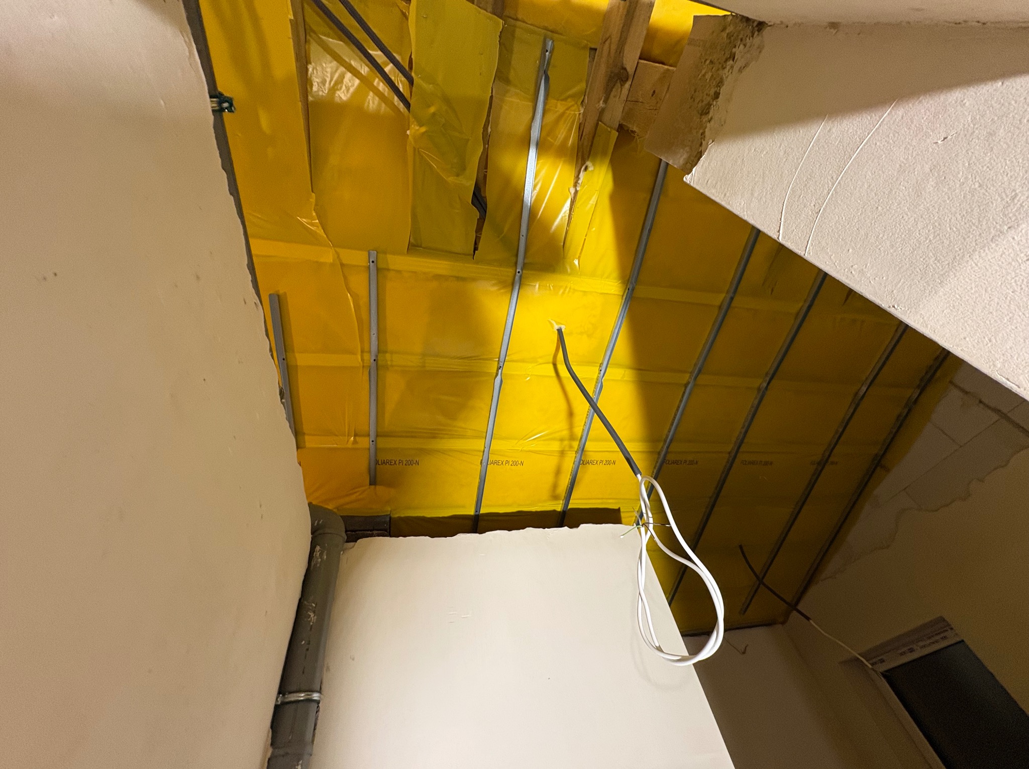 Żółta folia paroizolacyjna z metalowymi profilami na poddaszu, widok z dołu. Widoczne fragmenty ścian i wystające przewody elektryczne.