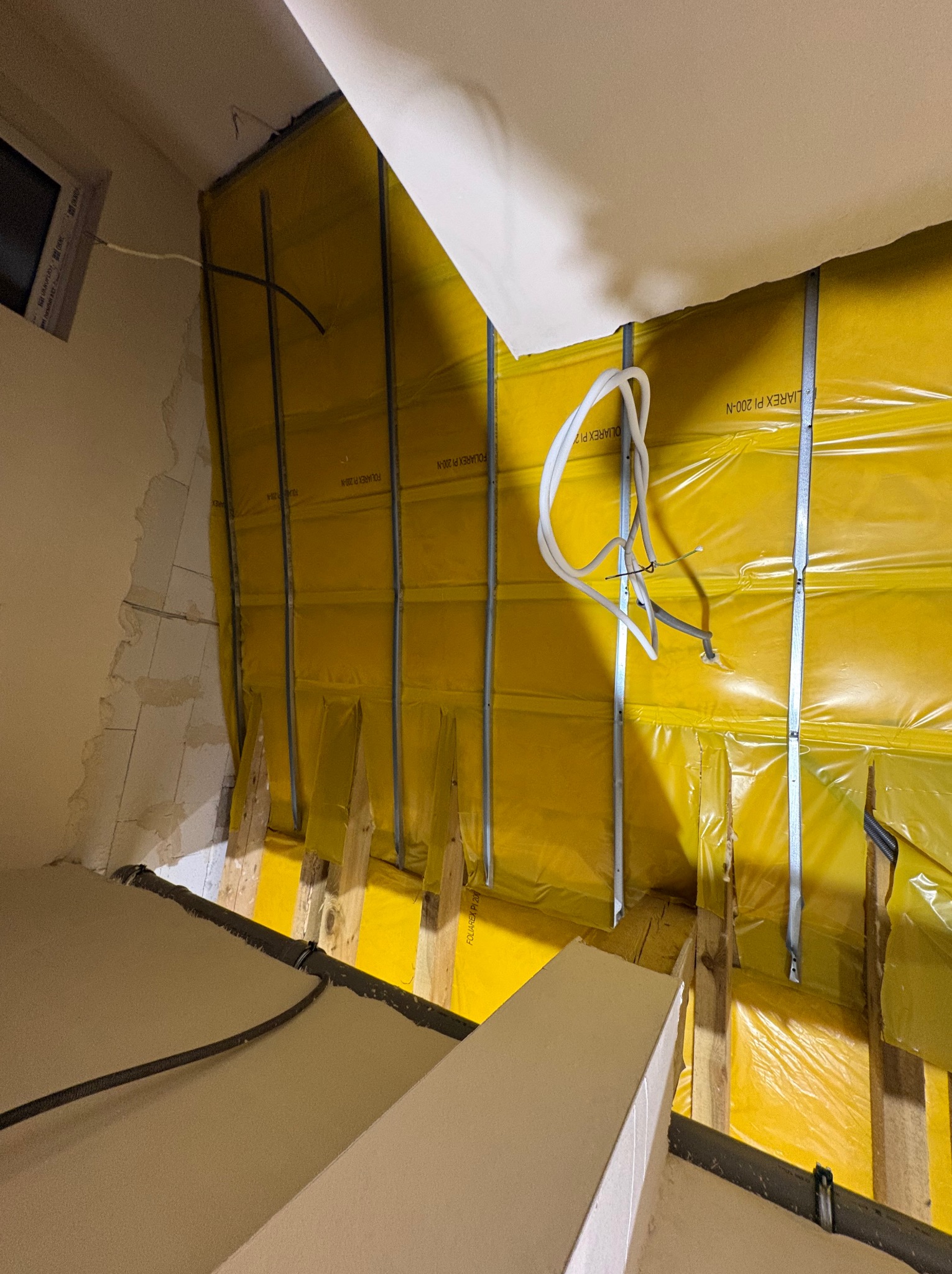 Poddasze w trakcie ocieplania: żółta folia paroizolacyjna, drewniane belki, metalowe profile, białe rury instalacyjne. Widoczne fragmenty ściany i sufitu.