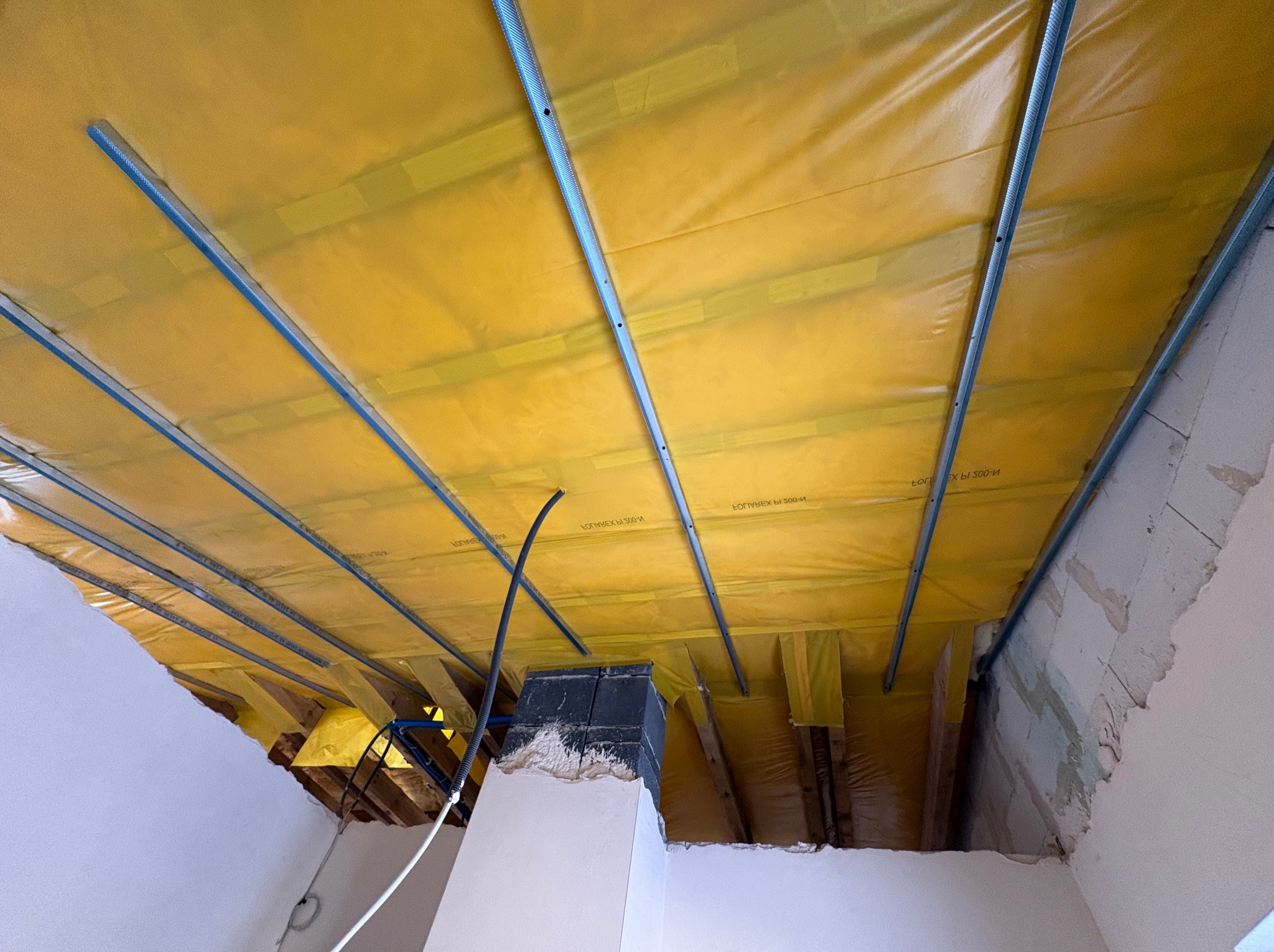 Żółta folia paroizolacyjna z niebieskimi profilami na poddaszu, widok z dołu. Fragment komina i ściany w surowym stanie. Etap prac wykończeniowych.