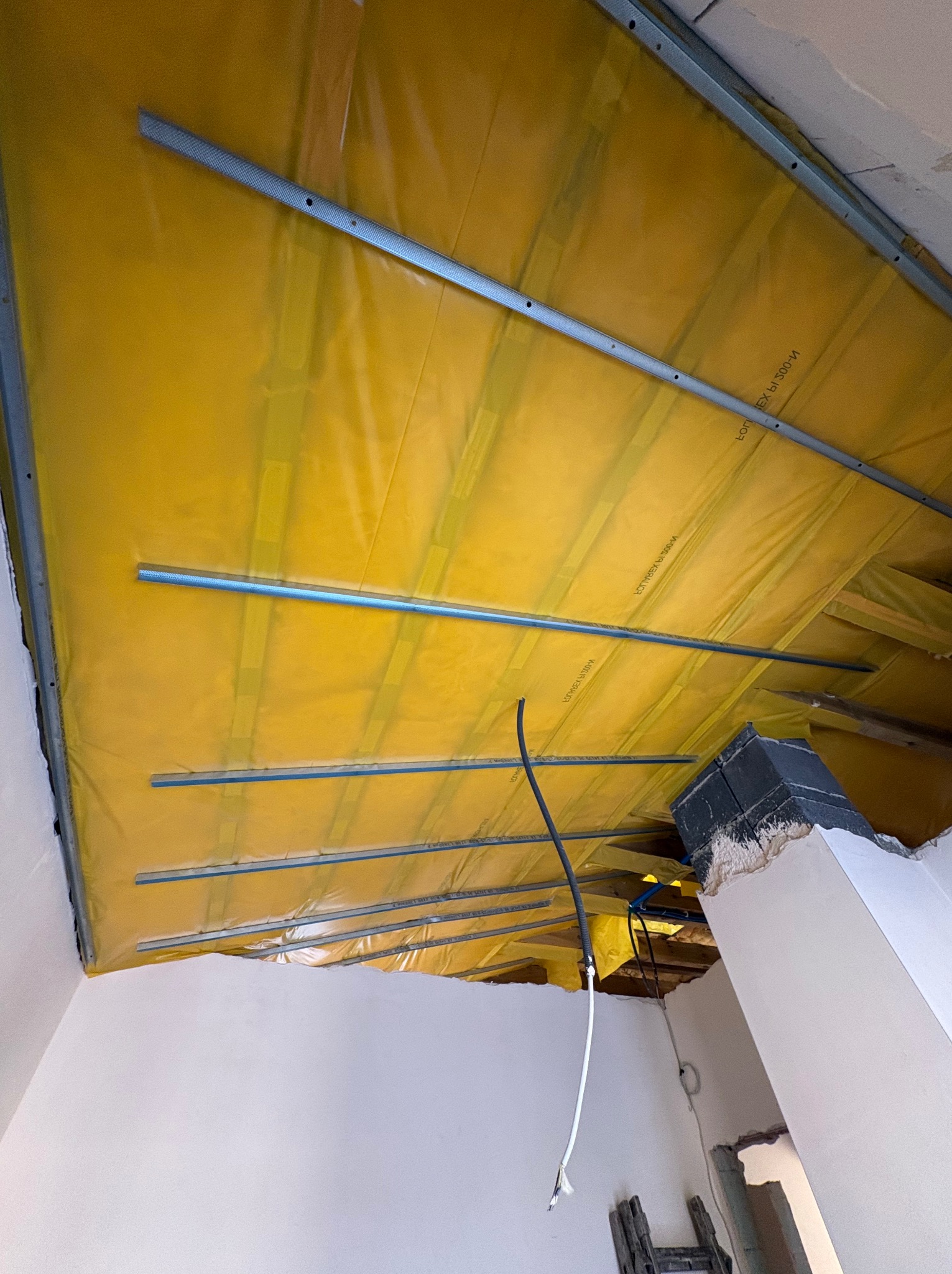 Żółta folia paroizolacyjna zamontowana na metalowym stelażu poddasza, widoczne przewody elektryczne i fragment komina. Prace wykończeniowe w trakcie.