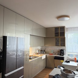Przemysław Kotowicz - Nowoczesna kuchnia z błyszczącymi frontami w odcieniach beżu i szarości, złote akcenty, sprzęt AGD, drewniana podłoga i okno z firanką. Całość doświetlona lampą sufitową.