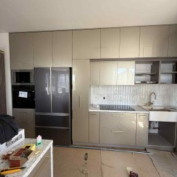 Przemysław Kotowicz - Beżowa kuchnia z połyskiem, zabudowa pod sufit z lodówką Haier, sprzęt AGD w zabudowie. Na blacie narzędzia i akcesoria monterskie. Podłoga zabezpieczona kartonem.