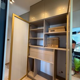 Przemysław Kotowicz - Otwarta szafa wnękowa z półkami i szufladami, wykończona w odcieniach beżu i złota. Widoczne kartony na półkach. Wnętrze oświetlone punktowo, drzwi z jasnego laminatu.