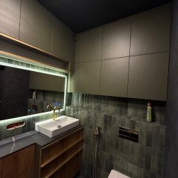 Przemysław Kotowicz - Nowoczesna łazienka z szafkami w odcieniach zieleni, podświetlanym lustrem i armaturą w kolorze złota, wykończona płytkami w odcieniach szarości.