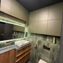 Nowoczesna łazienka z szafkami w odcieniach szarości i drewna, białą umywalką oraz złotą armaturą. Uwagę zwracają pionowe płytki w odcieniach szarości i czarny sufit.