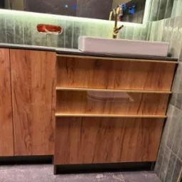 Drewniana szafka łazienkowa z umywalką na blacie, złota armatura, szare płytki w tle i podświetlane lustro. Widoczna instalacja elektryczna.