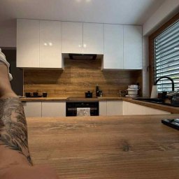 Przemysław Kotowicz - Nowoczesna kuchnia z białymi szafkami i drewnianym blatem, oświetlona czarnymi reflektorami. W tle okno z żaluzjami i fragment wytatuowanej ręki w kadrze.