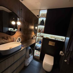 Przemysław Kotowicz - Nowoczesna łazienka z umywalką nablatową, szafką z szufladami, toaletą i półkami z oświetleniem LED. Ciemna kolorystyka, minimalistyczny design, nietypowe lustro.