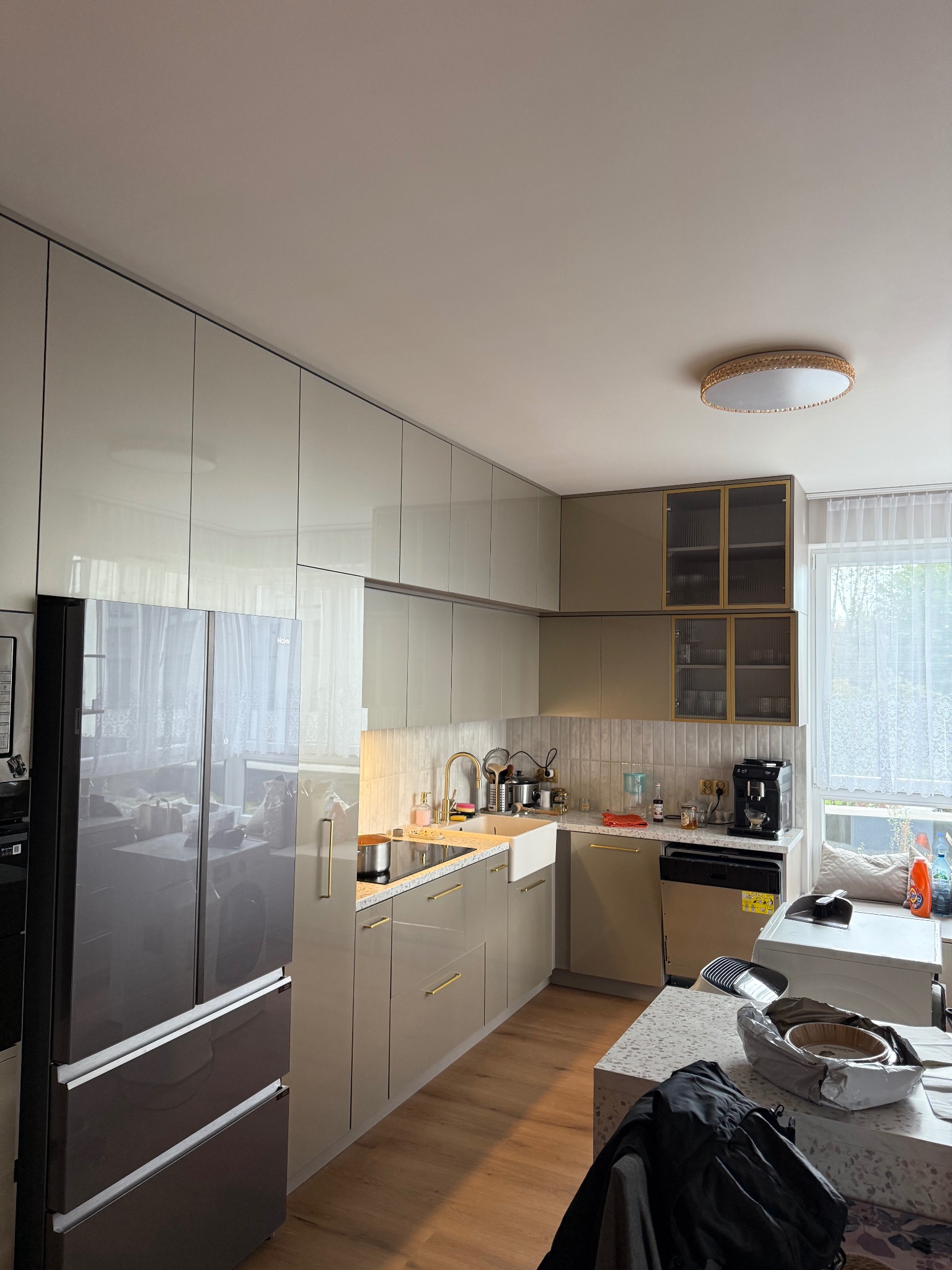 Nowoczesna kuchnia z błyszczącymi frontami w odcieniach beżu i szarości, złote akcenty, sprzęt AGD, drewniana podłoga i okno z firanką. Całość doświetlona lampą sufitową.