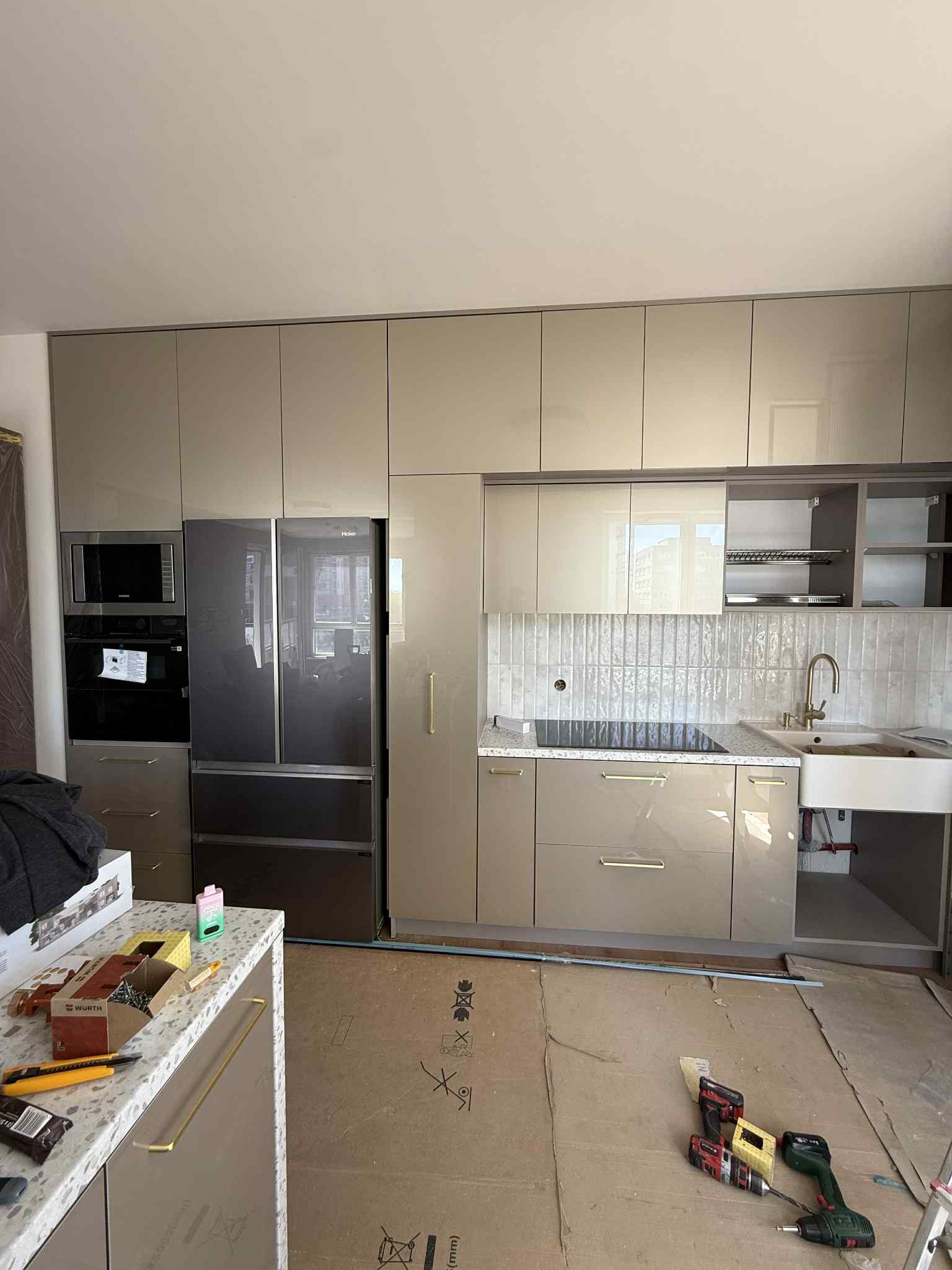Beżowa kuchnia z połyskiem, zabudowa pod sufit z lodówką Haier, sprzęt AGD w zabudowie. Na blacie narzędzia i akcesoria monterskie. Podłoga zabezpieczona kartonem.