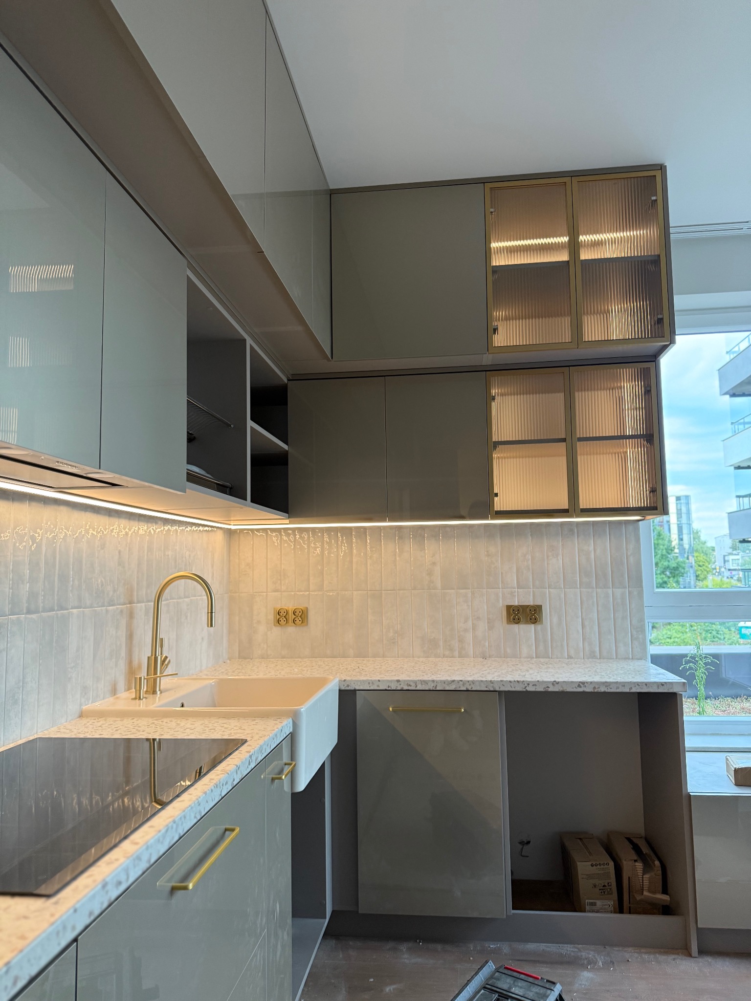 Nowoczesna kuchnia z szarymi, lakierowanymi frontami, złotymi detalami i oświetleniem LED. Widoczne szklane szafki górne i blat z konglomeratu.