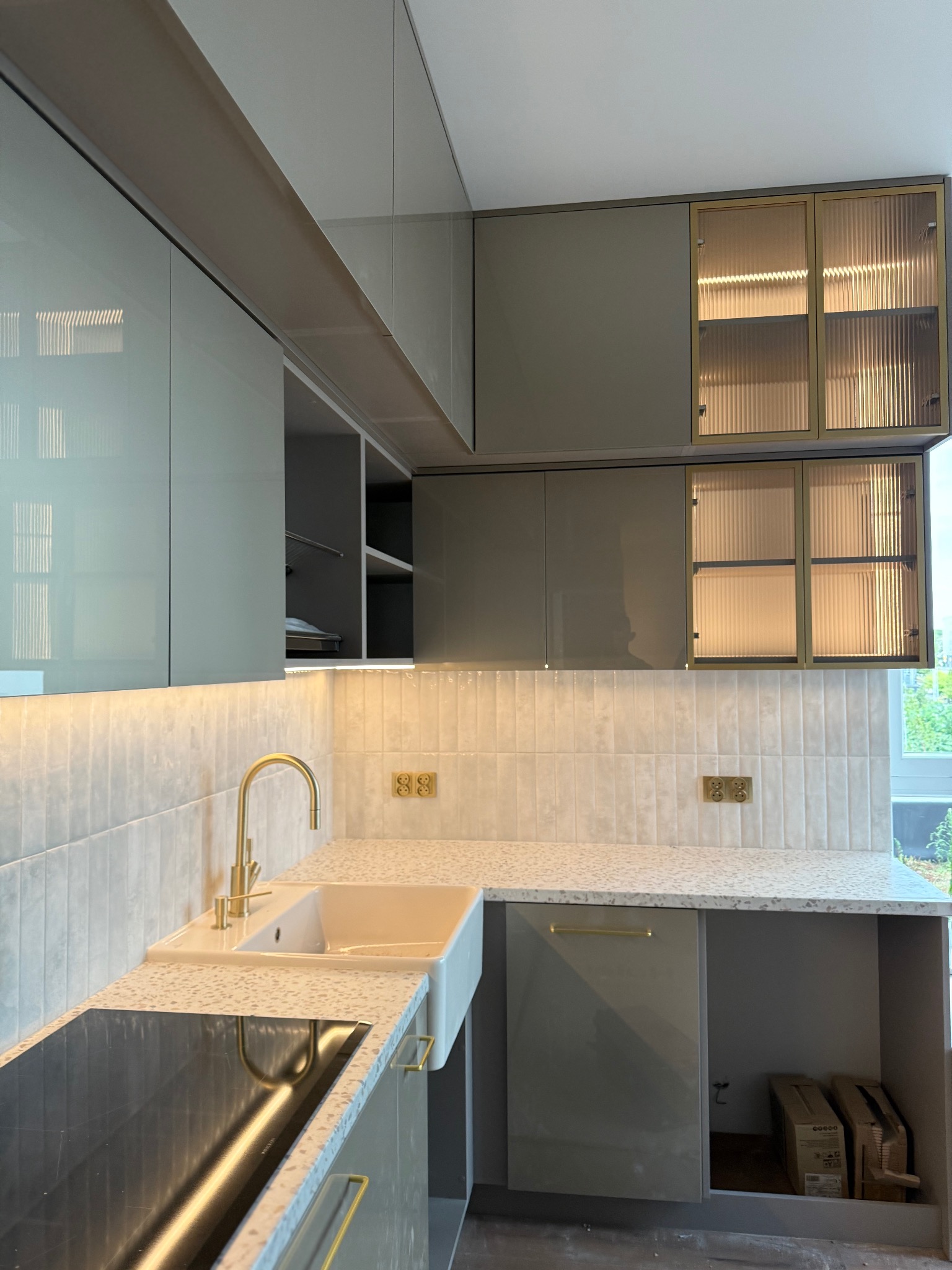 Nowoczesna kuchnia z szarymi szafkami, złotymi akcentami i białym blatem lastryko. Uwagę przykuwają ryflowane szklane fronty górnych szafek.