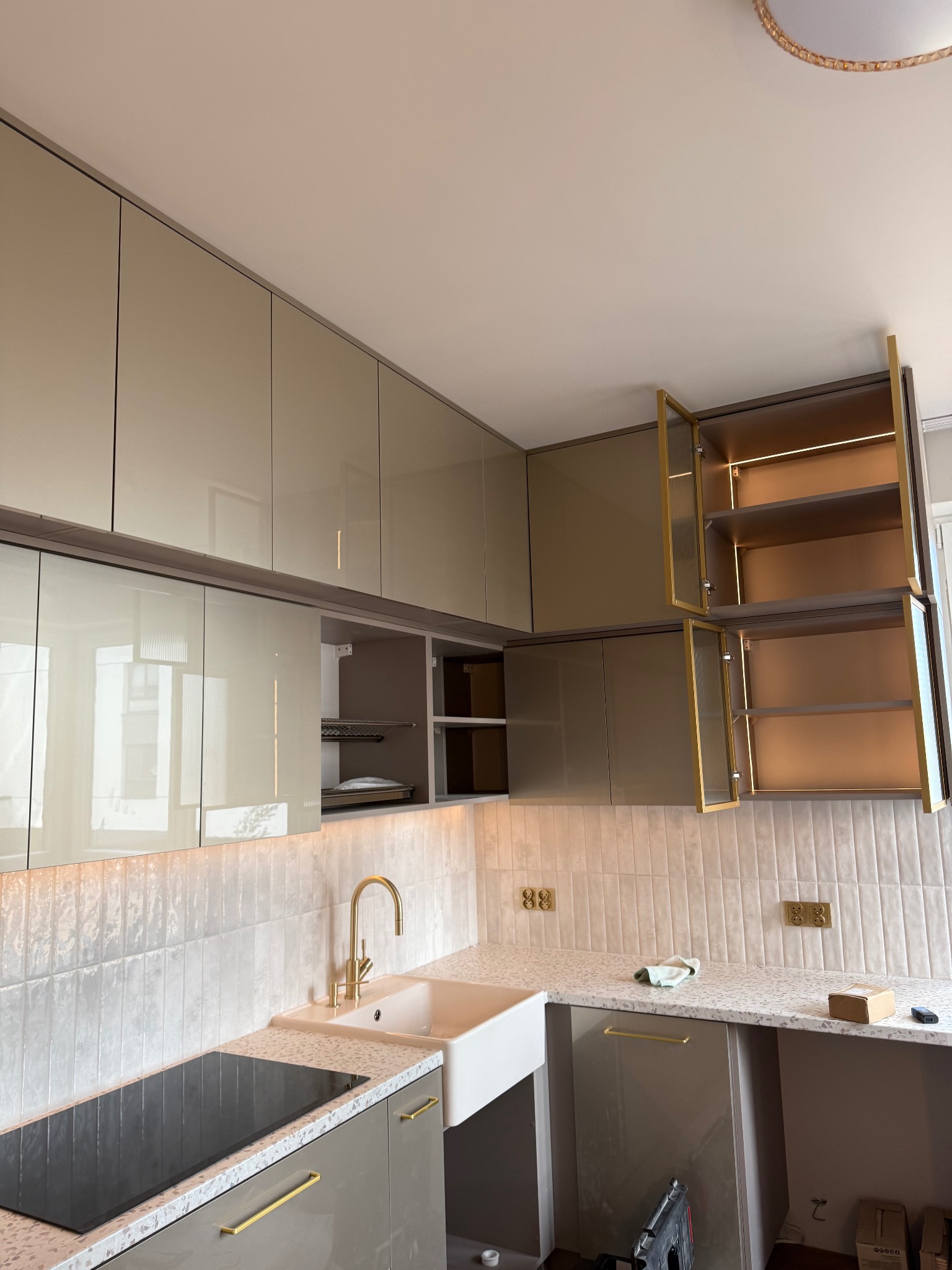 Beżowa kuchnia z górnymi szafkami z oświetleniem LED, złotymi uchwytami i baterią, białą umywalką, płytą indukcyjną i płytkami na ścianie.