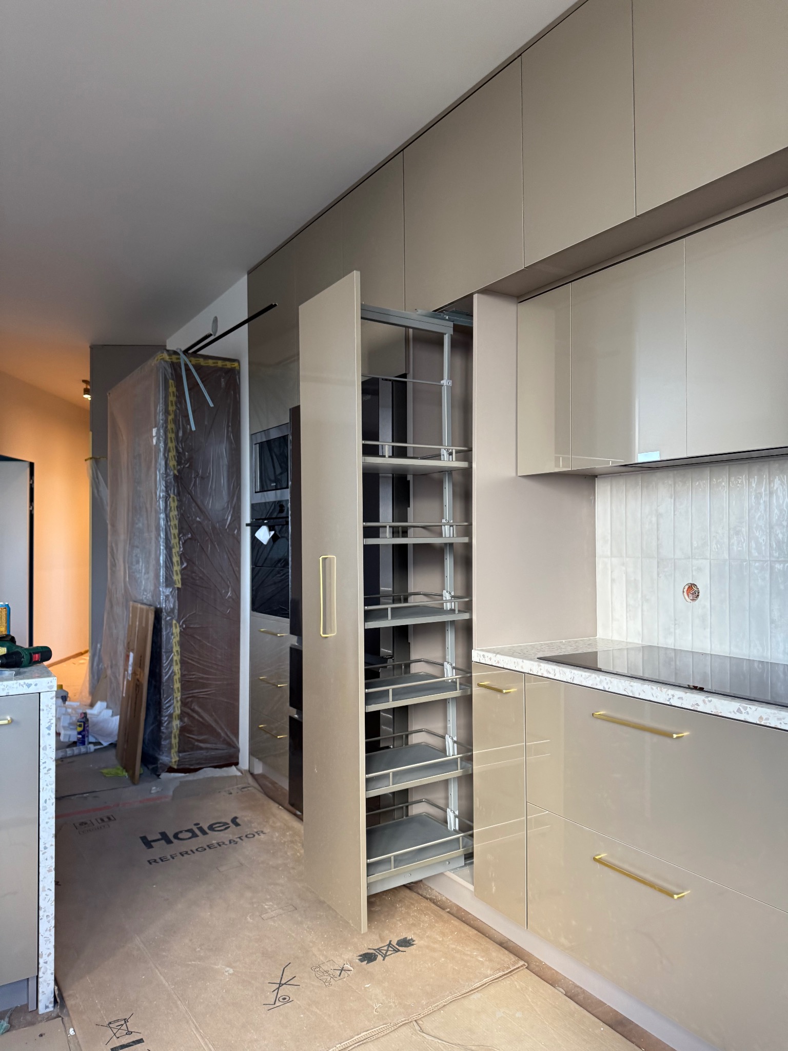 Beżowa kuchnia z wysuwaną szafką cargo i złotymi uchwytami. Podłoga zabezpieczona kartonem z logo Haier. W tle widoczny sprzęt AGD w zabudowie, zapakowany w folię.