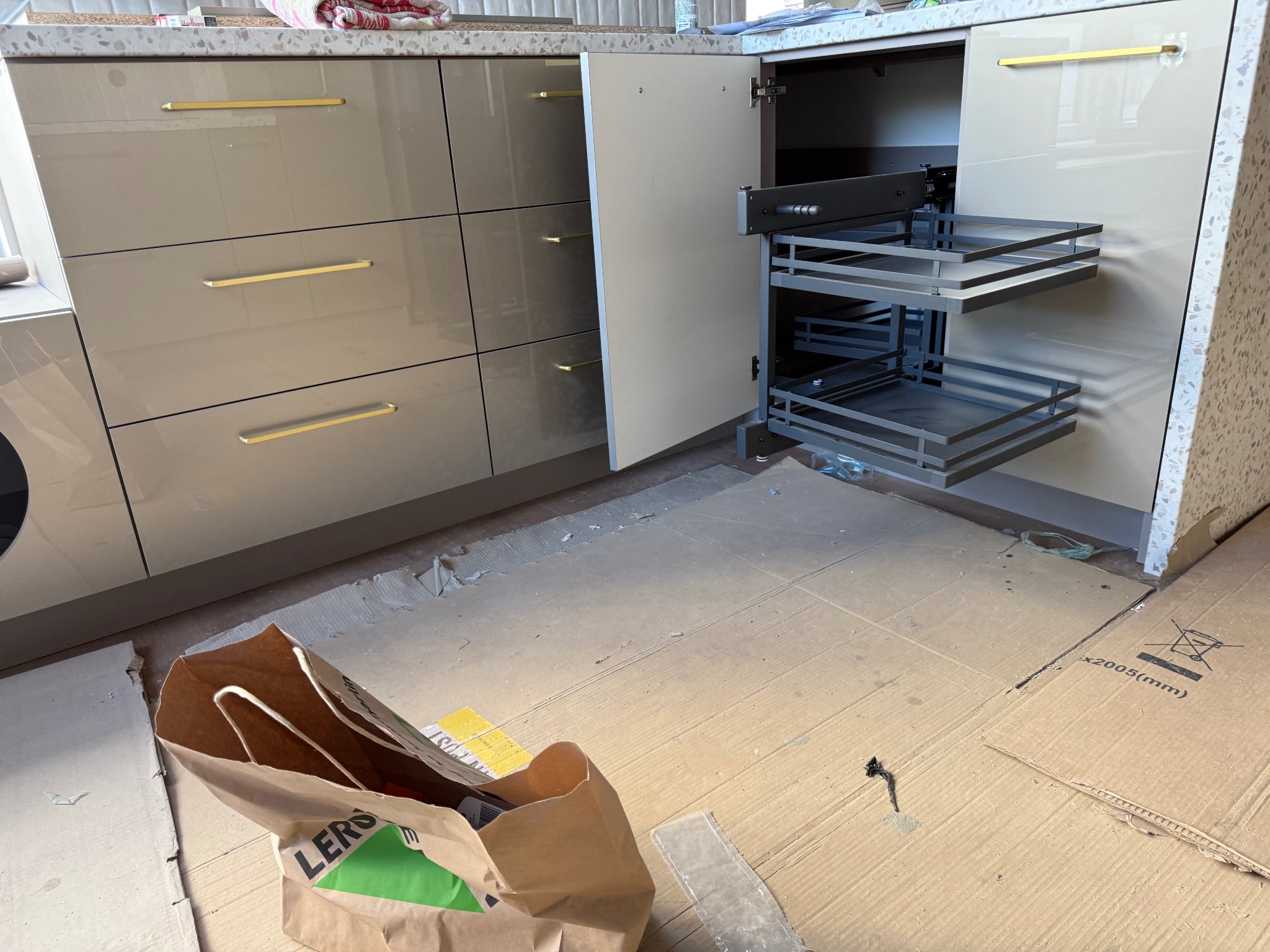 Beżowa kuchnia z wysuwanym koszem cargo. Widoczne złote uchwyty i blat z lastryko. Na podłodze kartony zabezpieczające.