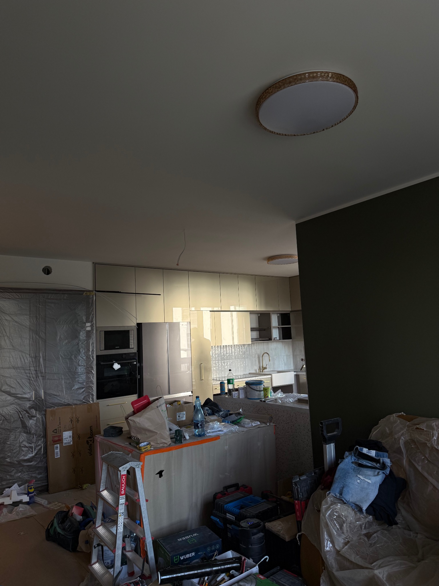 Montaż jasnej kuchni: widoczne szafki, wyspa kuchenna w trakcie instalacji, narzędzia i materiały budowlane. Folia ochronna na ścianie. W tle okno i oświetlenie sufitowe.