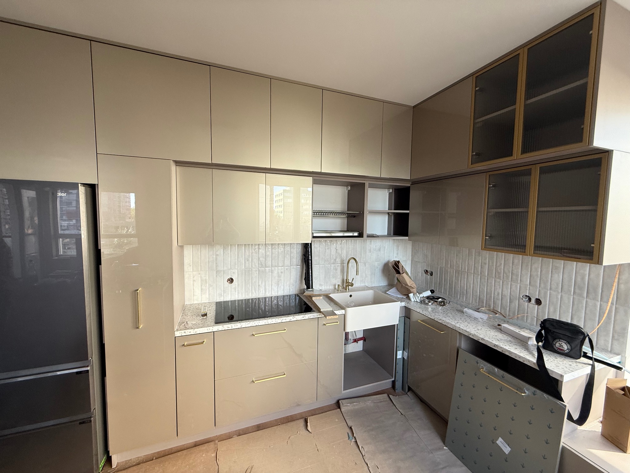 Beżowa kuchnia na wymiar z złotymi uchwytami i szklanymi szafkami wiszącymi, jeszcze w trakcie montażu, z widocznymi elementami wykończeniowymi i płytkami na ścianie.