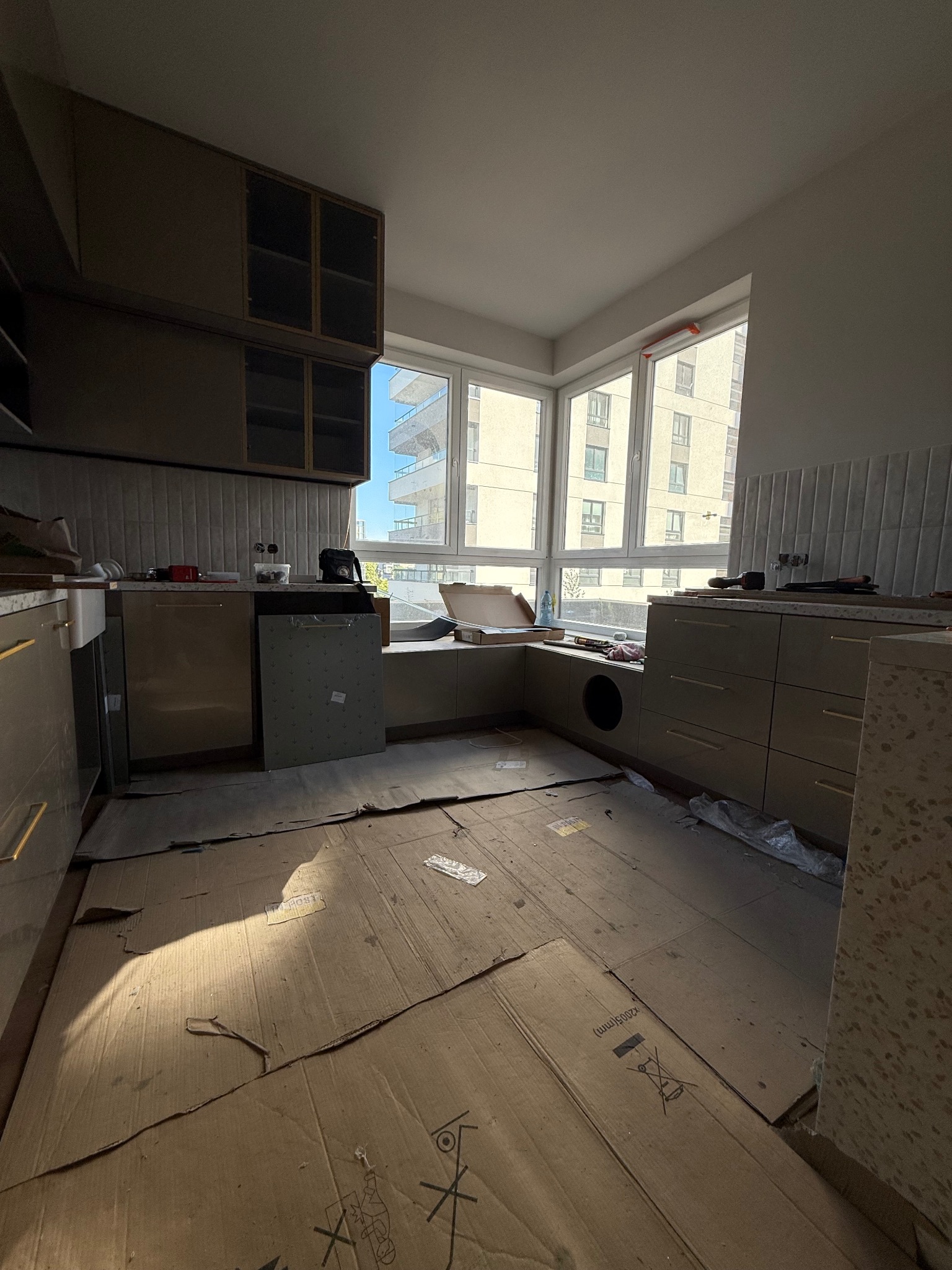 Montaż kuchni: beżowe szafki z złotymi uchwytami, okno z widokiem na budynek. Podłoga zabezpieczona kartonem, widoczne narzędzia i akcesoria montażowe.