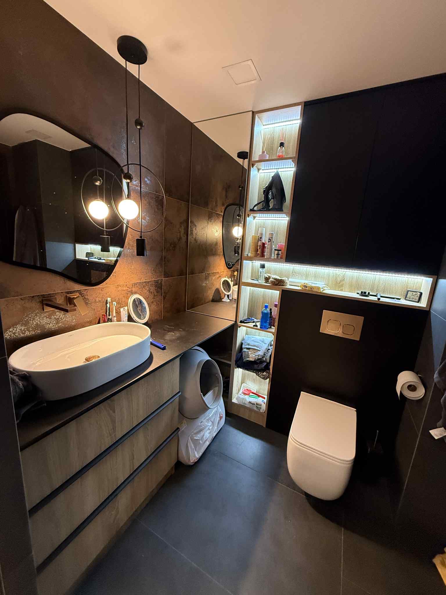 Nowoczesna łazienka z drewnianą szafką pod umywalką, lustrem o nieregularnym kształcie i oświetlonymi półkami. Ciemna kolorystyka, minimalistyczny design.