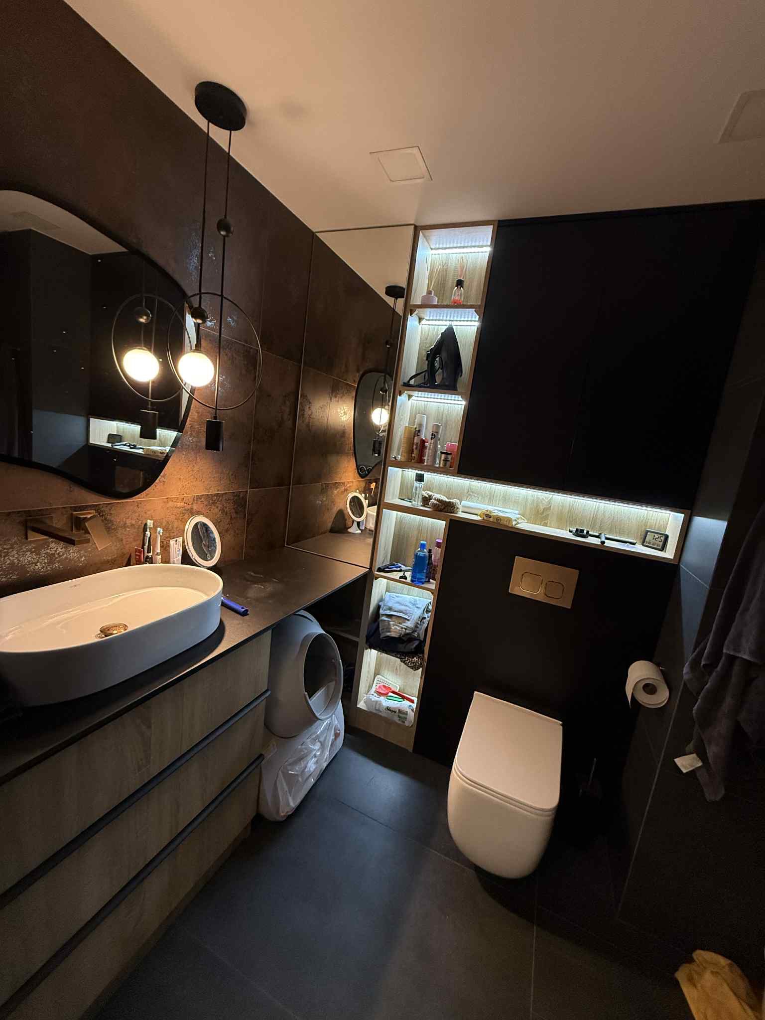 Nowoczesna łazienka z umywalką nablatową, szafką z szufladami, toaletą i półkami z oświetleniem LED. Ciemna kolorystyka, minimalistyczny design, nietypowe lustro.