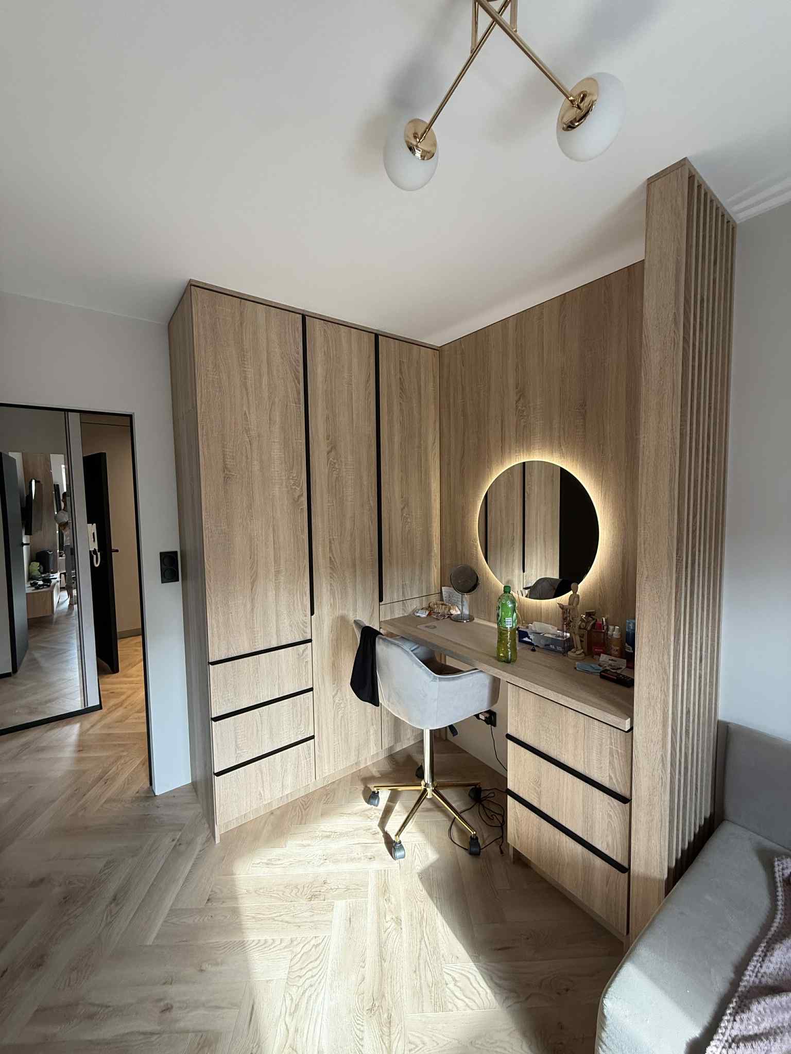 Jasna garderoba z toaletką i okrągłym lustrem z podświetleniem LED. Drewnopodobne fronty z czarnymi uchwytami. Parkiet w jodełkę. Nowoczesne oświetlenie sufitowe.