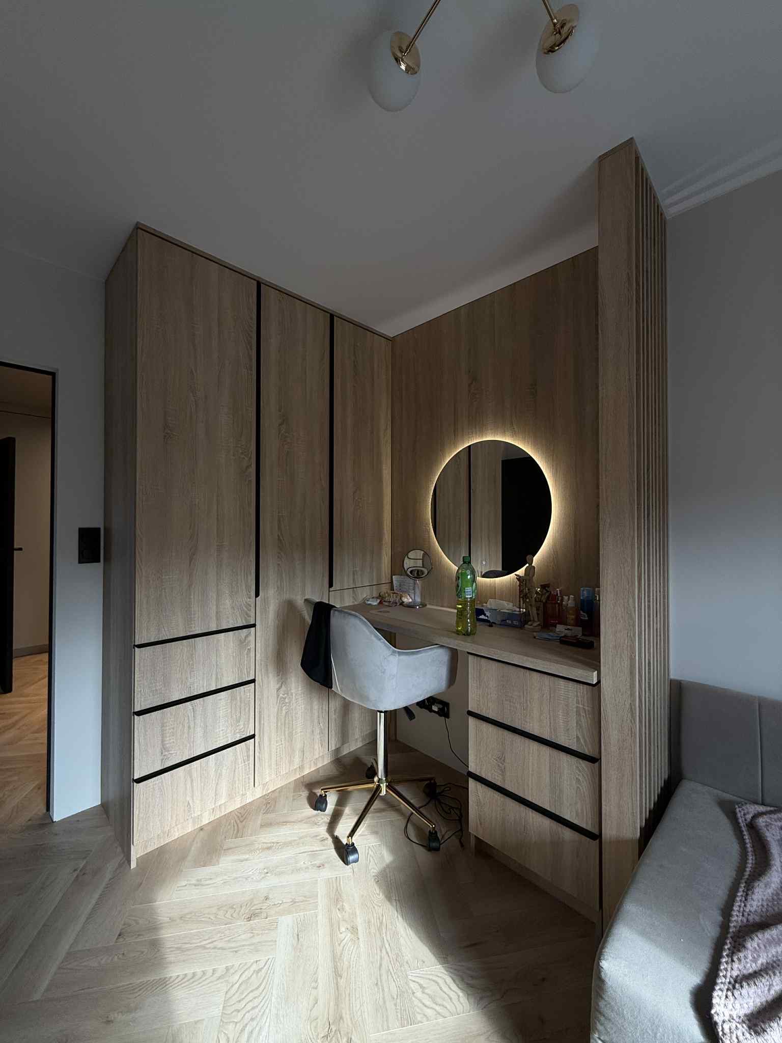 Jasna, drewniana szafa z czarnymi uchwytami i toaletka z okrągłym lustrem LED. Krzesło na złotych kółkach, parkiet w jodełkę. Nowoczesny design.