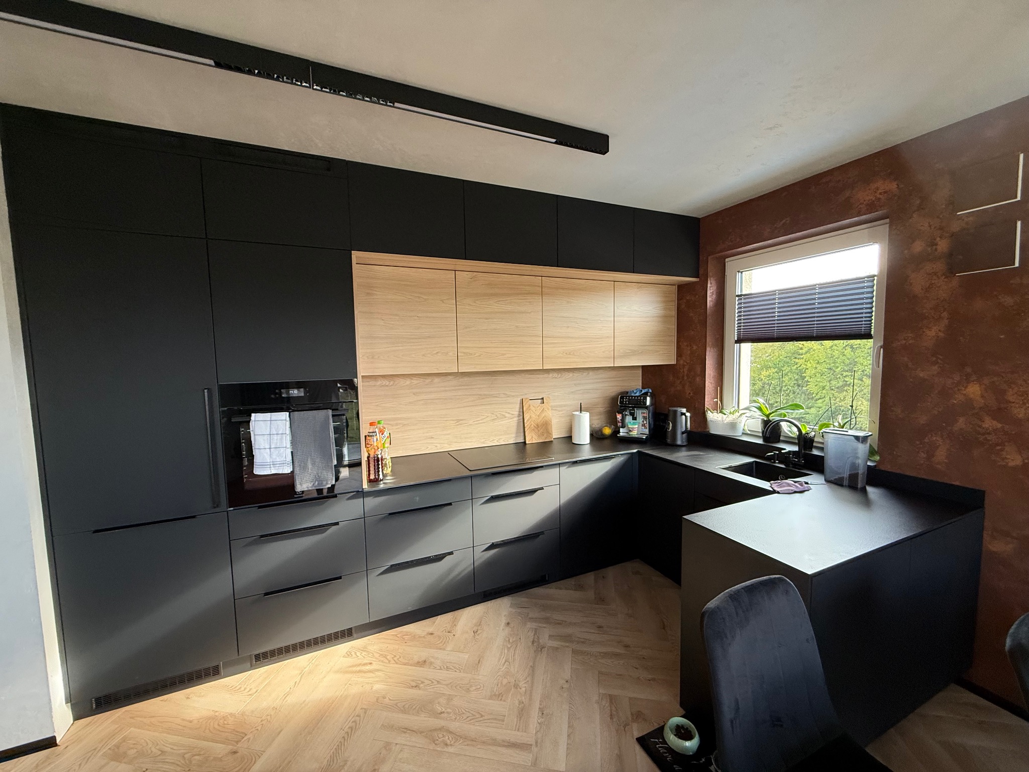 Nowoczesna kuchnia na wymiar z czarnymi frontami i drewnianymi szafkami wiszącymi, oświetlona listwą LED, z blatem roboczym i parkietem w jodełkę.