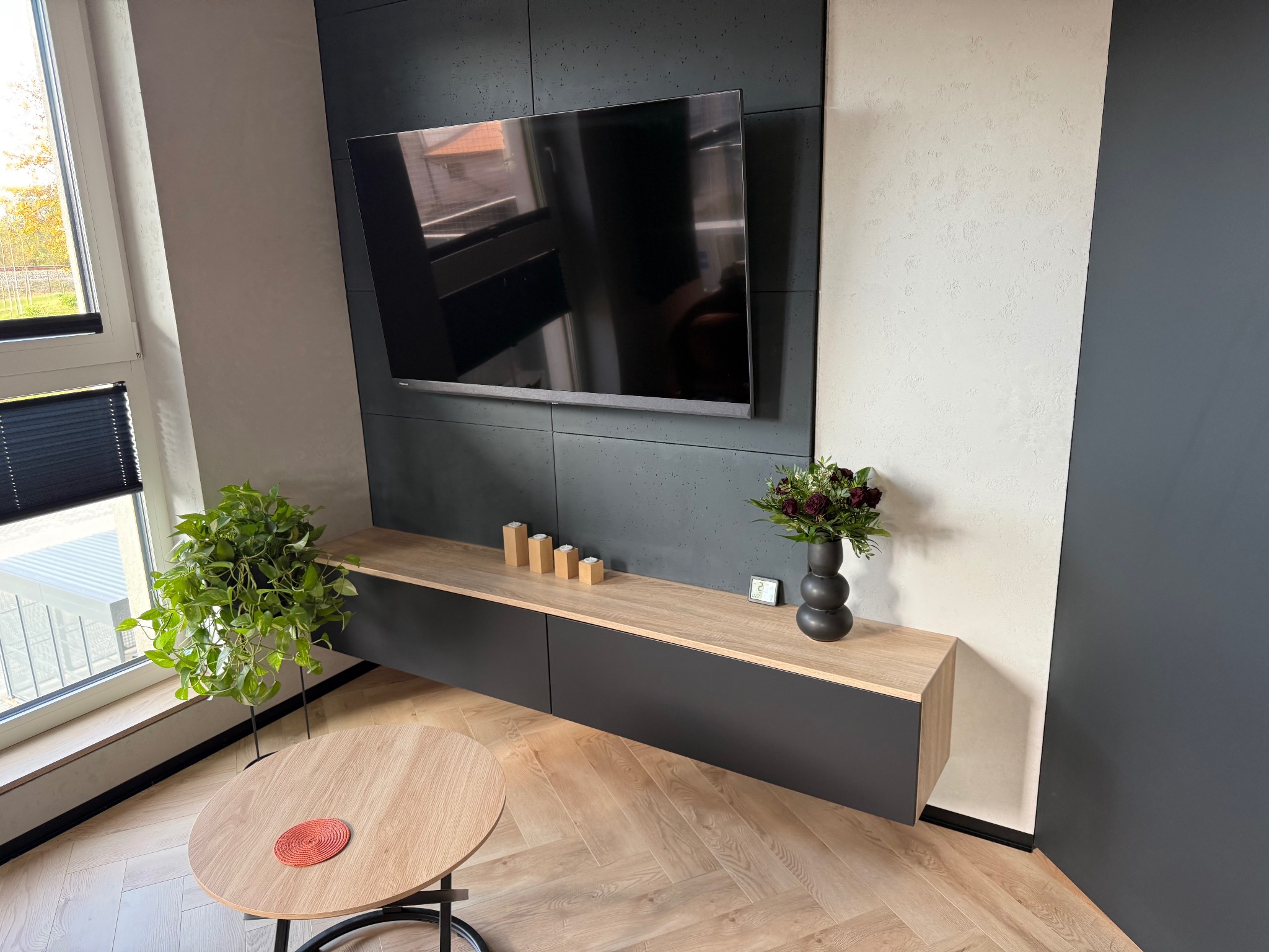 Nowoczesny salon z szafką RTV w kolorze dębu i grafitu, wiszącą pod telewizorem. Na blacie dekoracje: świeczki, zegar i wazon z kwiatami. Parkiet w jodełkę.