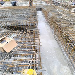 AG Budownictwo Firma Budowlana - Zbrojenie fundamentów budynku w trakcie prac. Widoczne stalowe pręty tworzące siatkę zbrojeniową, częściowo zalane betonem. Na pierwszym planie kartonowe pudełko i narzędzia.