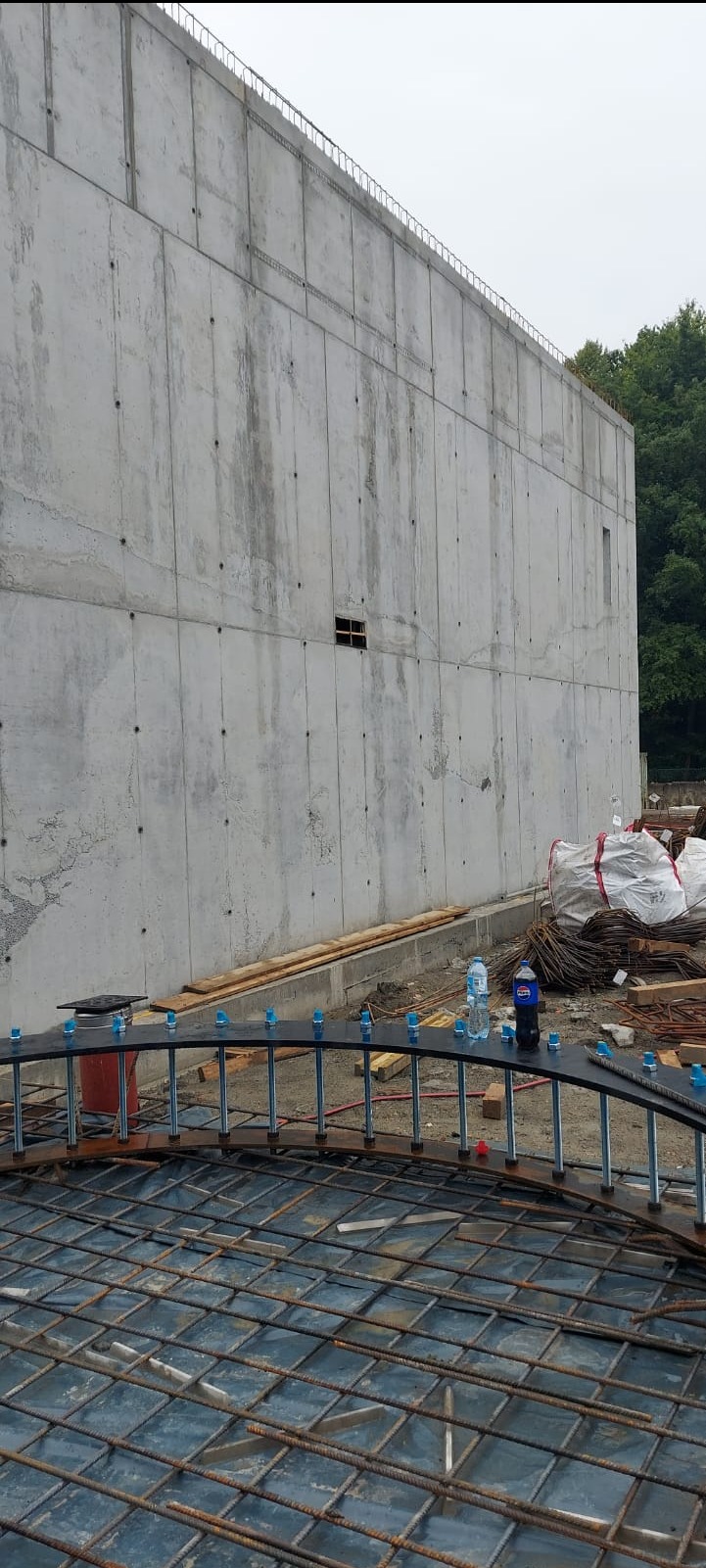 Ściana żelbetowa w surowym stanie z widocznym zbrojeniem w dolnej części kadru. Na zbrojeniu stoją dwie butelki wody i puszka napoju.