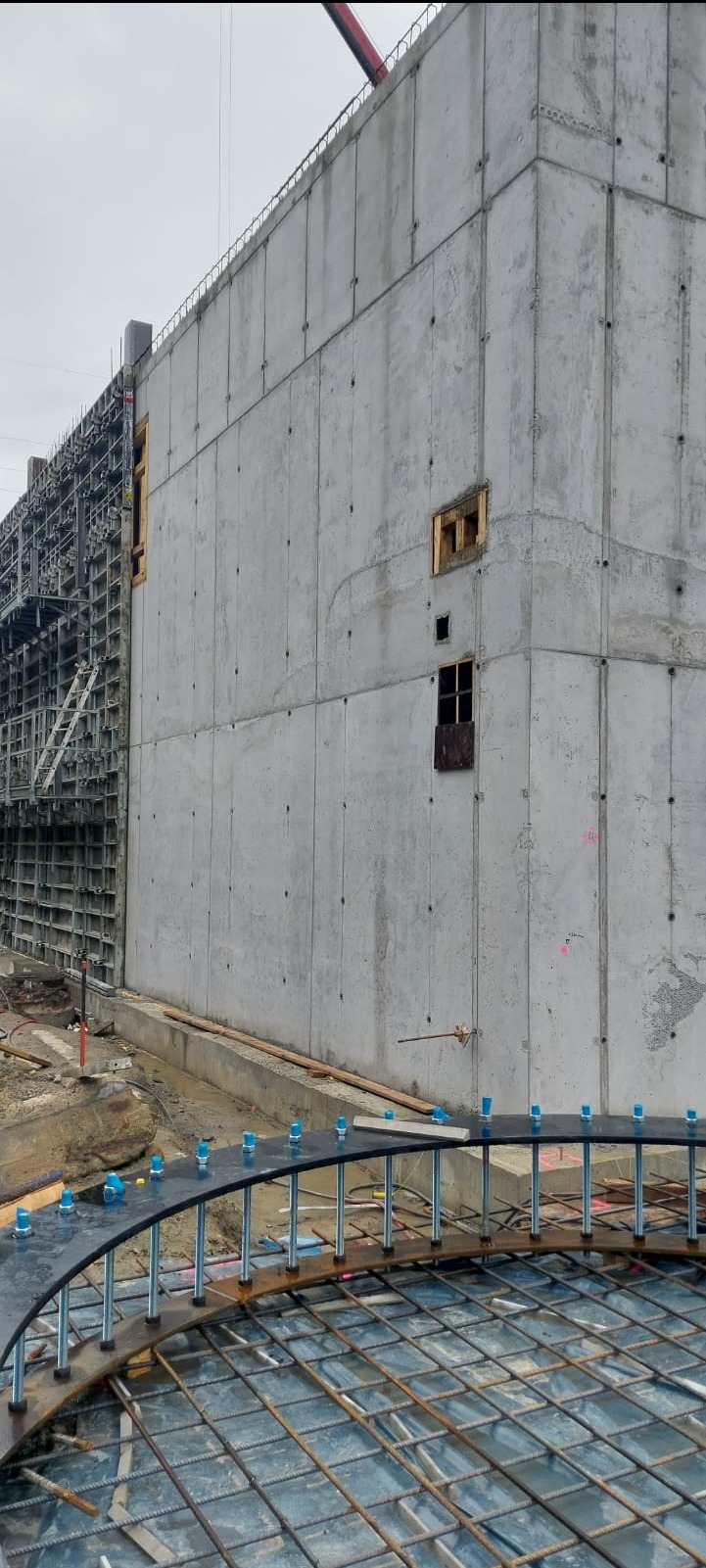 Betonowa ściana budynku w trakcie budowy z otworami okiennymi i widocznym zbrojeniem, uchwycona w pochmurny dzień z bliska.