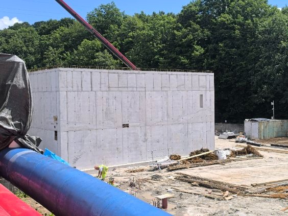 Żelbetowa konstrukcja budynku w trakcie budowy, widoczna szalowana ściana z betonu, rura do pompowania betonu i pracownik w oddali. W tle zieleń drzew.