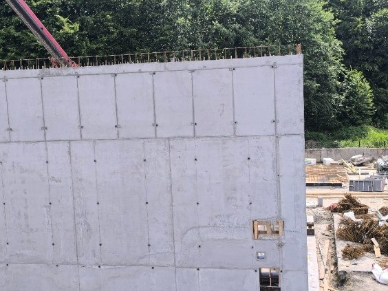 Żelbetowa ściana budynku w trakcie budowy, widoczne zbrojenia i fragment żurawia. Tło stanowią drzewa i elementy placu budowy.