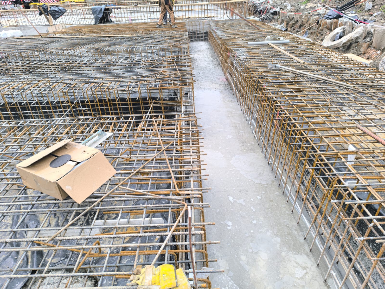 Zbrojenie fundamentów budynku w trakcie prac. Widoczne stalowe pręty tworzące siatkę zbrojeniową, częściowo zalane betonem. Na pierwszym planie kartonowe pudełko i narzędzia.