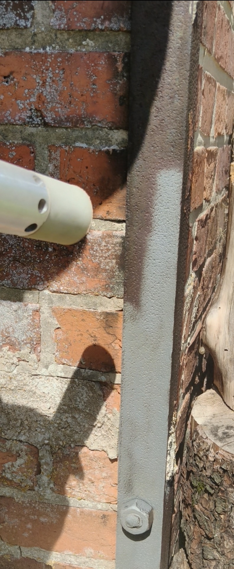 Ceglana ściana z fragmentami porostów czyszczona punktowo urządzeniem z dyszą, obok metalowa konstrukcja po czyszczeniu, na pniu drzewa mały ślimak.