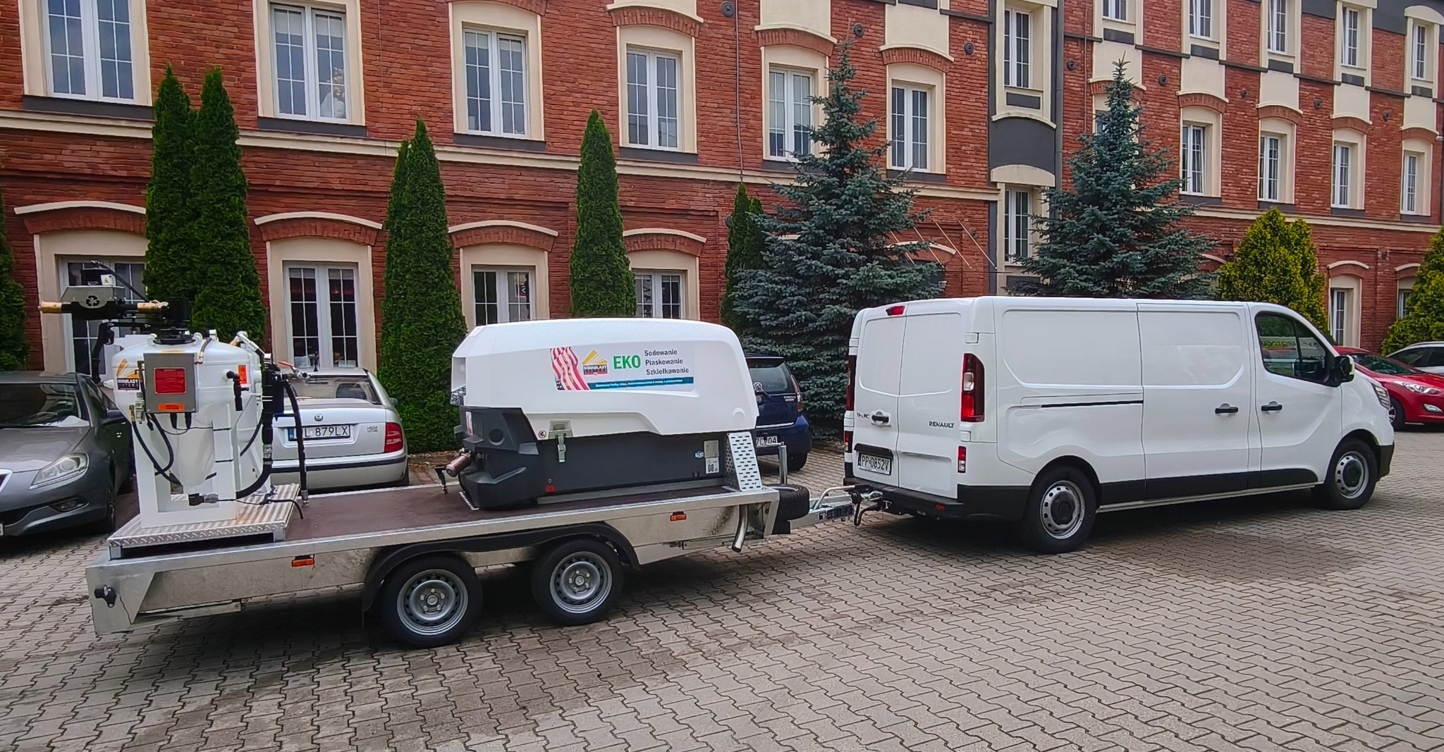 Mobilna piaskarka EKO na przyczepie, ciągnięta przez biały samochód dostawczy Renault na tle ceglanego budynku z zielenią. Sprzęt do sodowania, piaskowania i szkiełkowania.