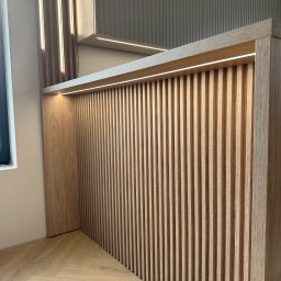 KS Meble - Nowoczesna zabudowa meblowa z ryflowanym frontem i oświetleniem LED, wykonana z drewna w jasnym odcieniu dębu, z widocznym fragmentem podłogi w jodełkę.