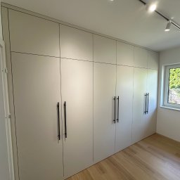 KS Meble - Jasna szafa wnękowa z czarnymi, minimalistycznymi uchwytami, wkomponowana w przestrzeń pokoju z drewnianą podłogą i oświetleniem punktowym.