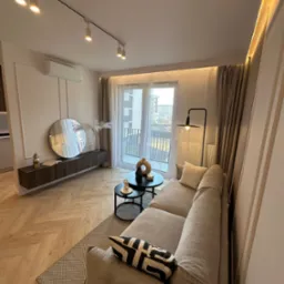 Salon w Frydrychowicach z szafką RTV, okrągłym lustrem, sofą i stolikami kawowymi. Parkiet ułożony w jodełkę. Nowoczesne oświetlenie sufitowe.