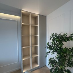 KS Meble - Wysoka, beżowa szafa wnękowa z oświetleniem LED, wkomponowana w ścianę z dekoracyjnymi listwami. Obok stoi doniczkowa roślina, a w kadrze widoczny fragment fotela.