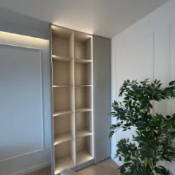 Wysoka, beżowa szafa wnękowa z oświetleniem LED, wkomponowana w ścianę z dekoracyjnymi listwami. Obok stoi doniczkowa roślina, a w kadrze widoczny fragment fotela.