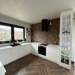 KS Meble - Nowoczesna kuchnia z białymi frontami, drewnianą ścianą dekoracyjną i podłogą w jodełkę. Na blacie przy oknie widoczne rośliny i akcesoria kuchenne.