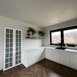 KS Meble - Jasna kuchnia z białymi szafkami, drewnianą podłogą w jodełkę i oknem z widokiem na zieleń. Na półce rośliny doniczkowe i dekoracje.